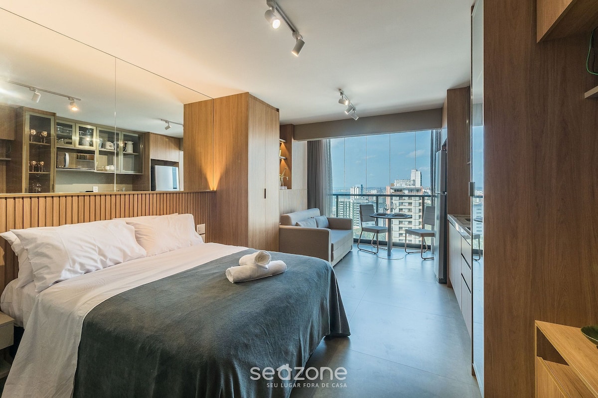 A modern hotel room with a bed, a seating area, a view of the city, and a kitchenette.