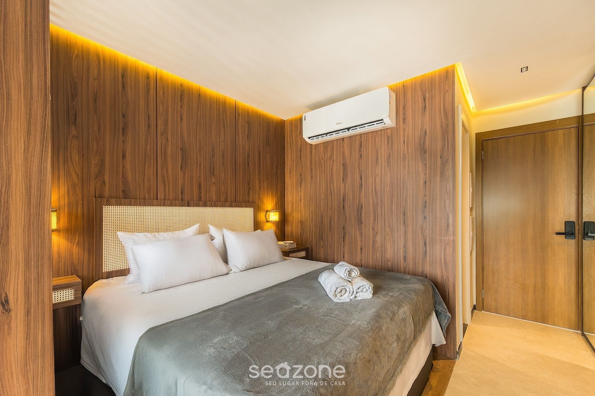A hotel room with a wooden panel wall, a bed with white bedding, a wall-mounted air conditioner, and a wooden wardrobe.