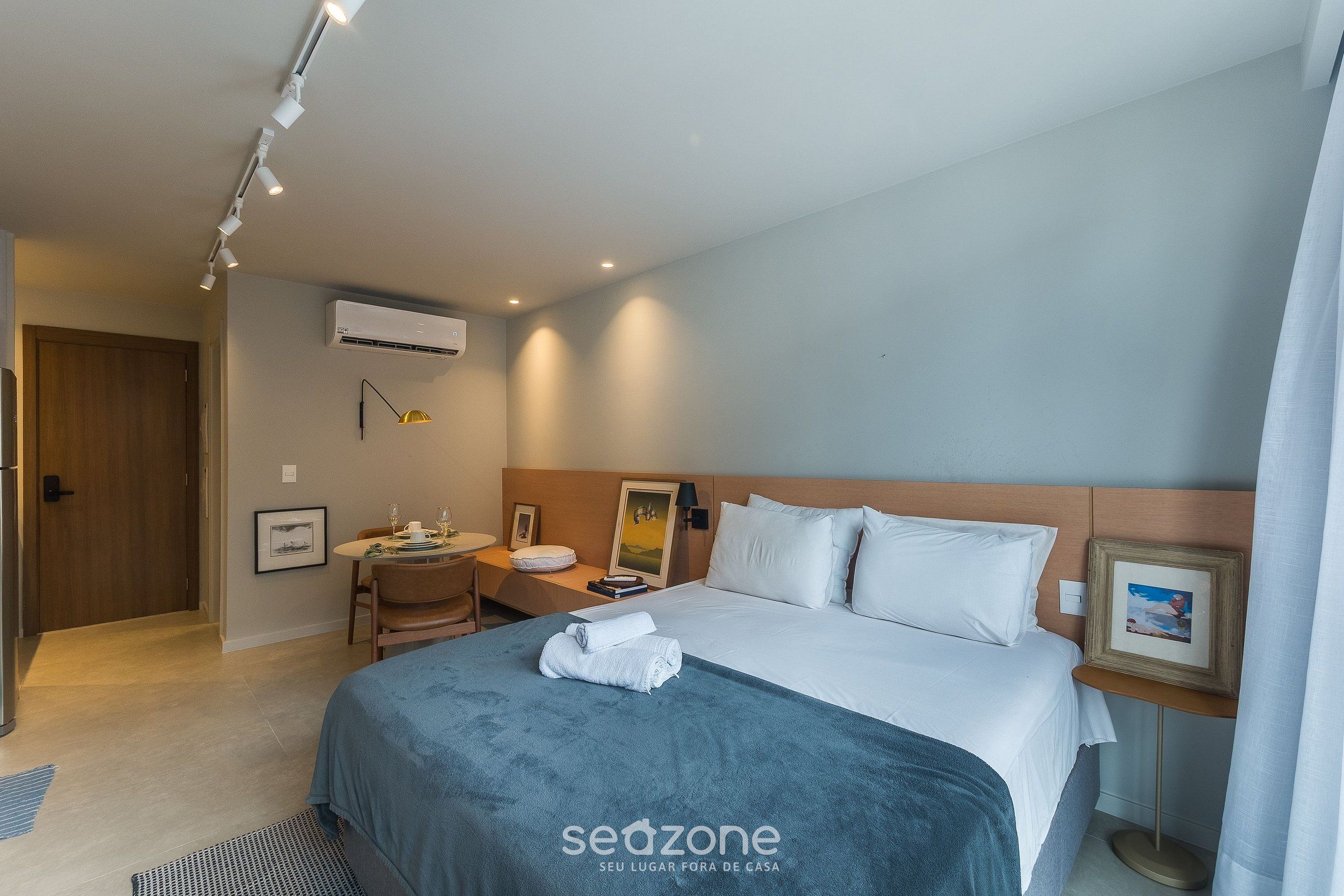 A hotel room with a bed, bedside table, lamp, framed picture, and an air conditioning unit.