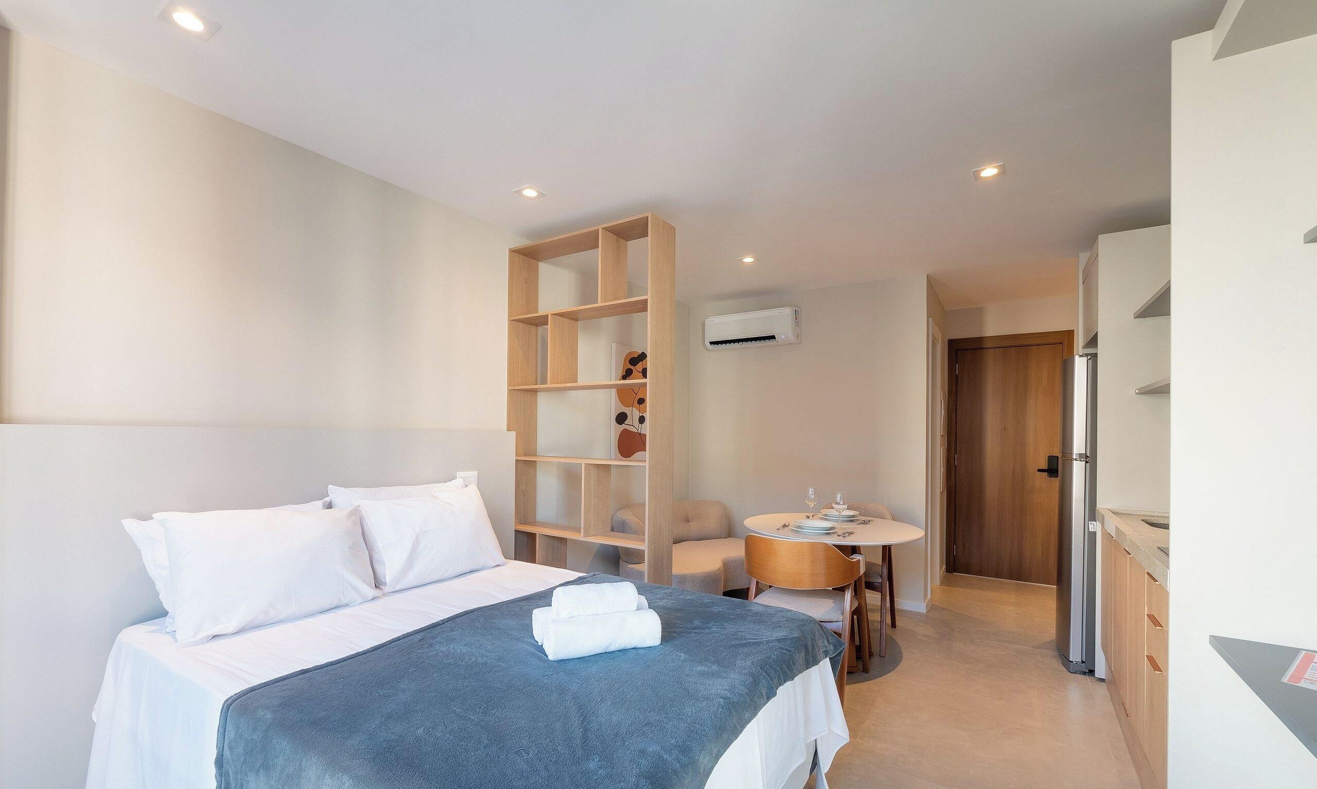 Habitación de hotel con cama, una pequeña mesa de comedor con sillas, una estantería de madera y una kitchenette.