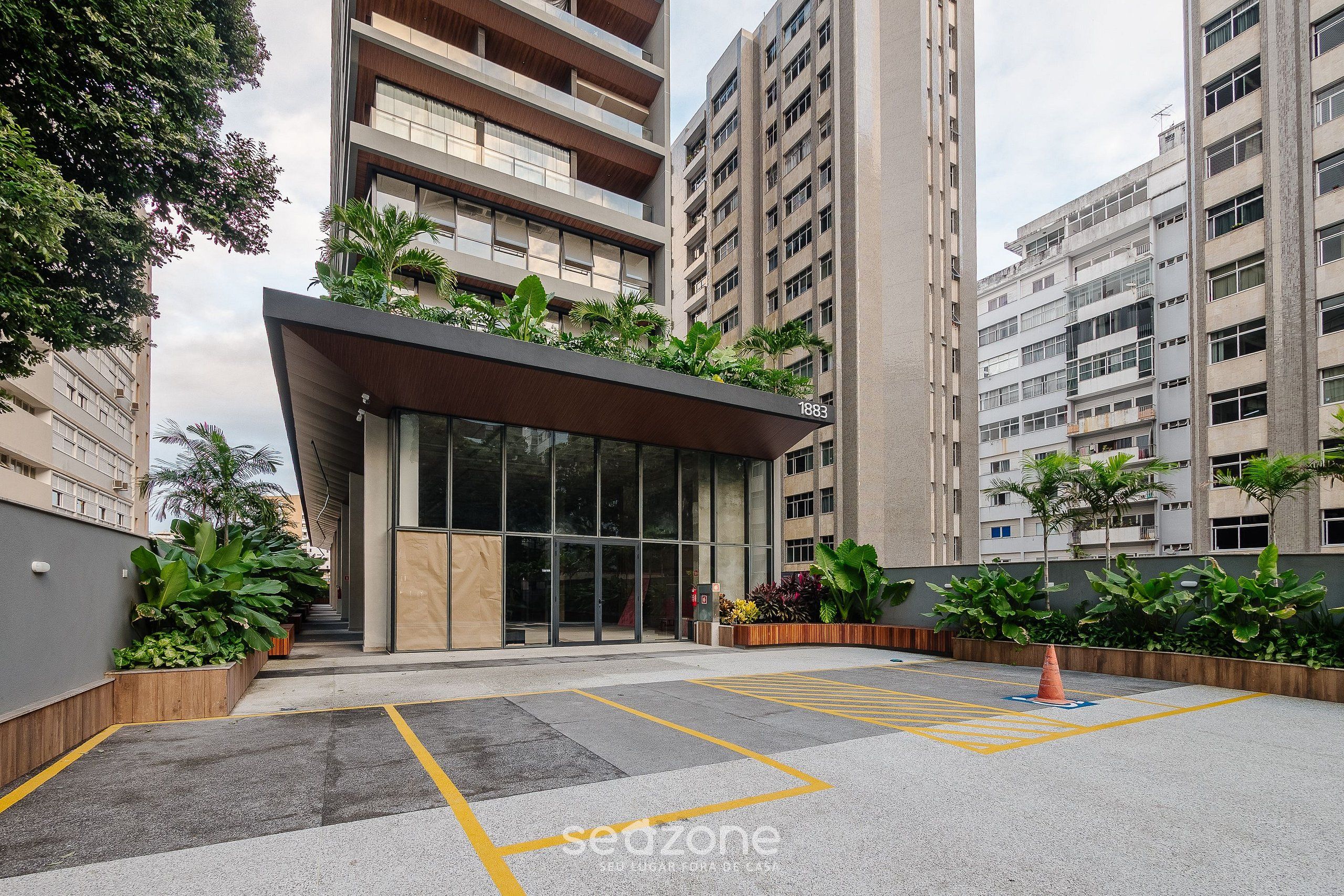 Ingresso moderno di un edificio con facciata in vetro, una pensilina e un'area di parcheggio delimitata da linee gialle.