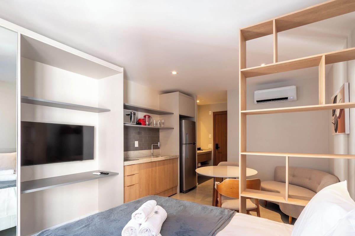 A modern hotel room with a bed, a small kitchenette, a dining area, and a built-in TV.