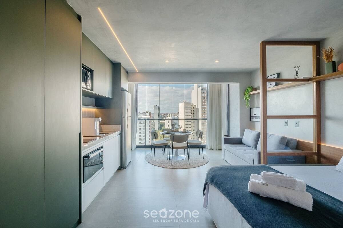 A modern hotel room with a kitchen, dining area, and a balcony with a city view.