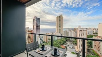 Un balcone con due tavoli, ognuno con una tazza e un cucchiaino, affaccia su una città con edifici alti e vista sul mare.