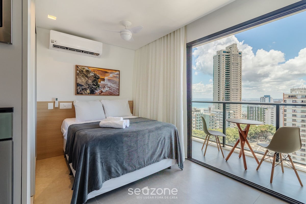 A hotel room with a bed, a painting, a balcony with chairs, and a view of a cityscape.