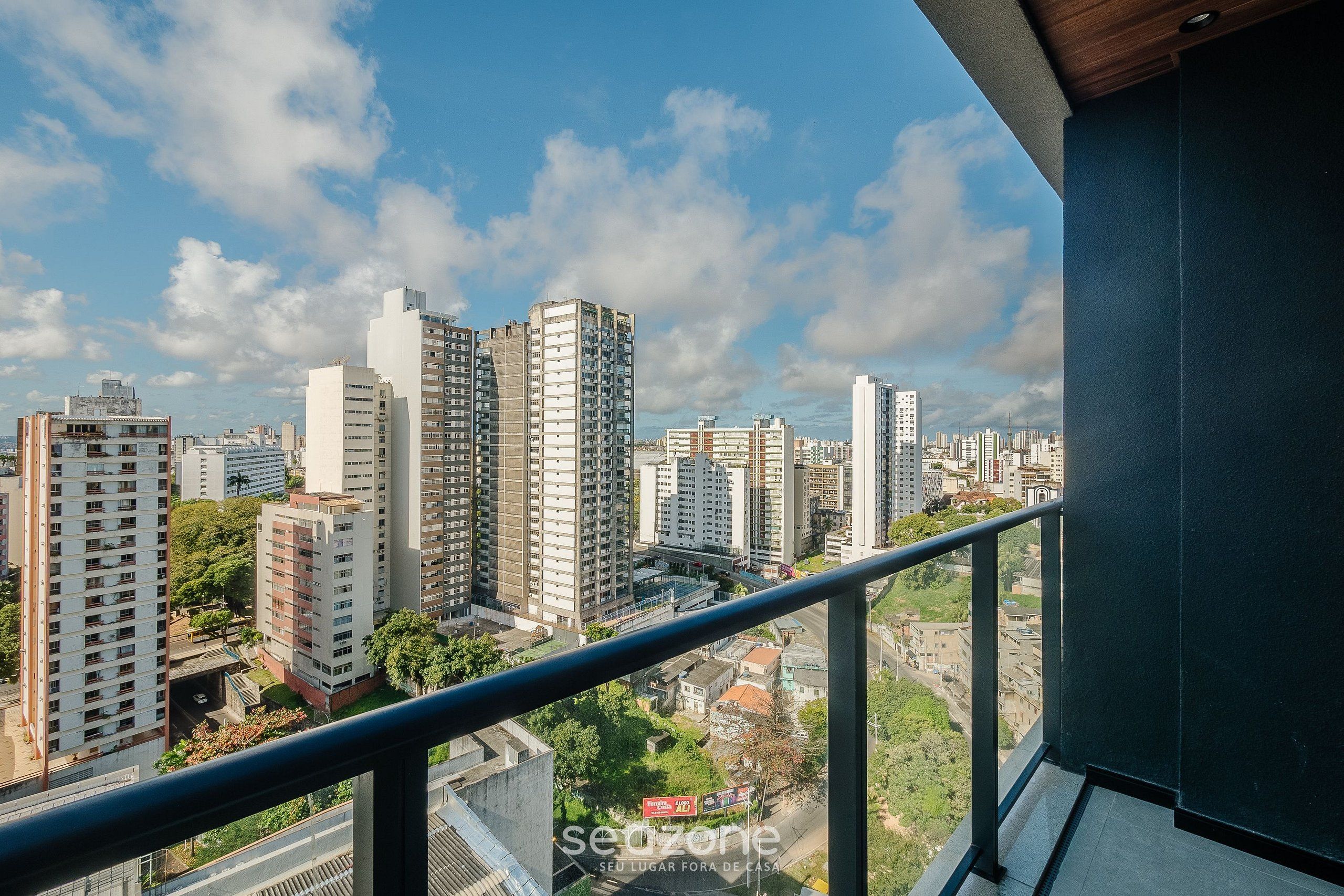 A high-rise building with multiple floors, a balcony with a metal railing, and a clear sky with clouds.