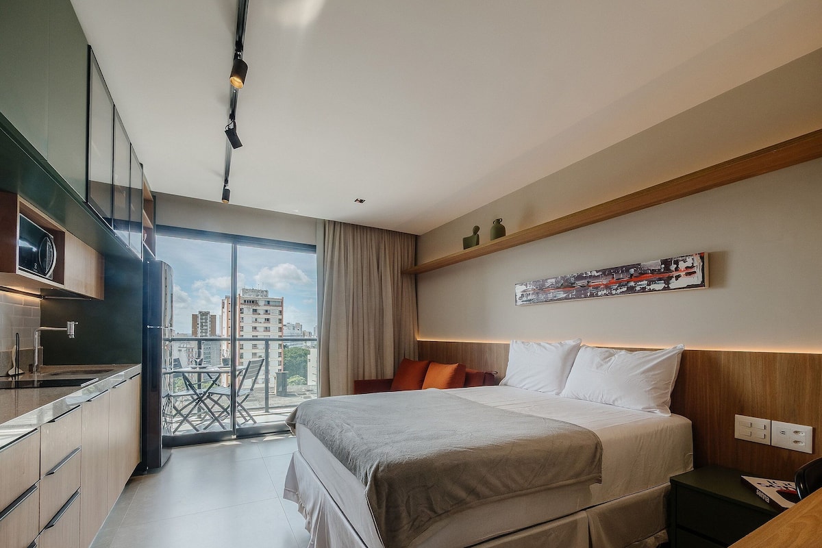 A modern hotel room with a bed, a small desk, a kitchenette, and a balcony with a city view.