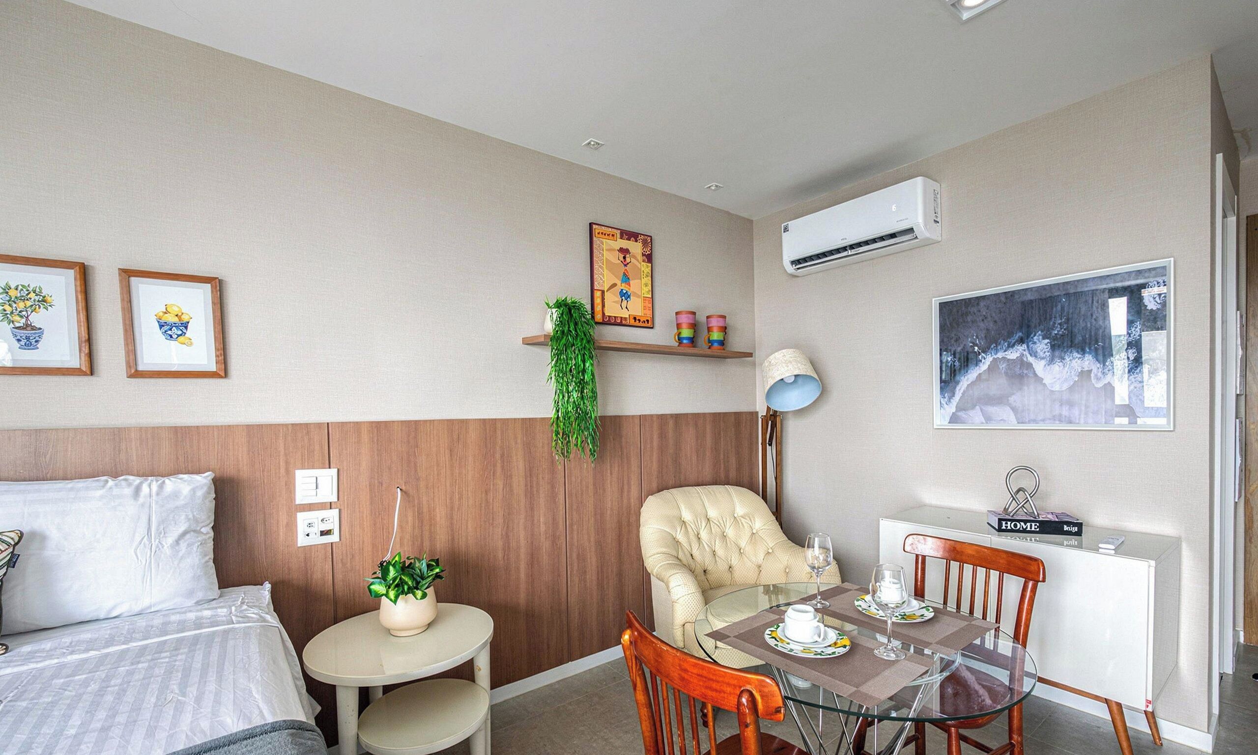Habitación de hotel con una cama, una pequeña mesa redonda con una silla, una mesa de comedor con sillas, una planta en maceta, un aire acondicionado montado en la pared y un cuadro enmarcado.
