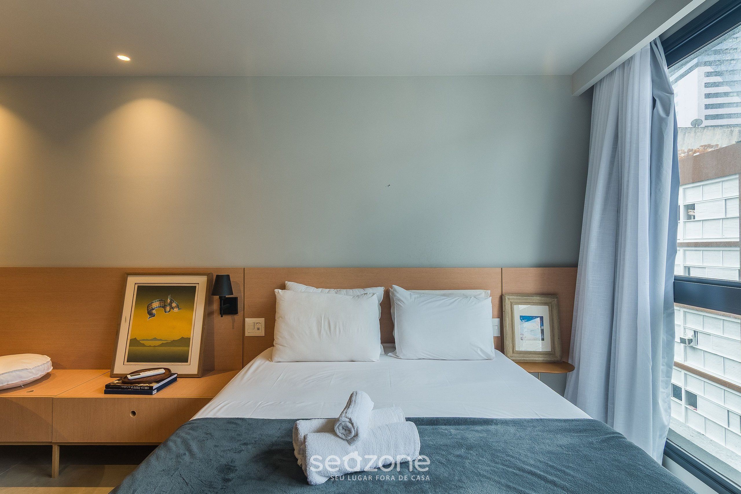 Une chambre d’hôtel avec un lit, une table de chevet, un tableau encadré, une serviette et une fenêtre avec des rideaux.