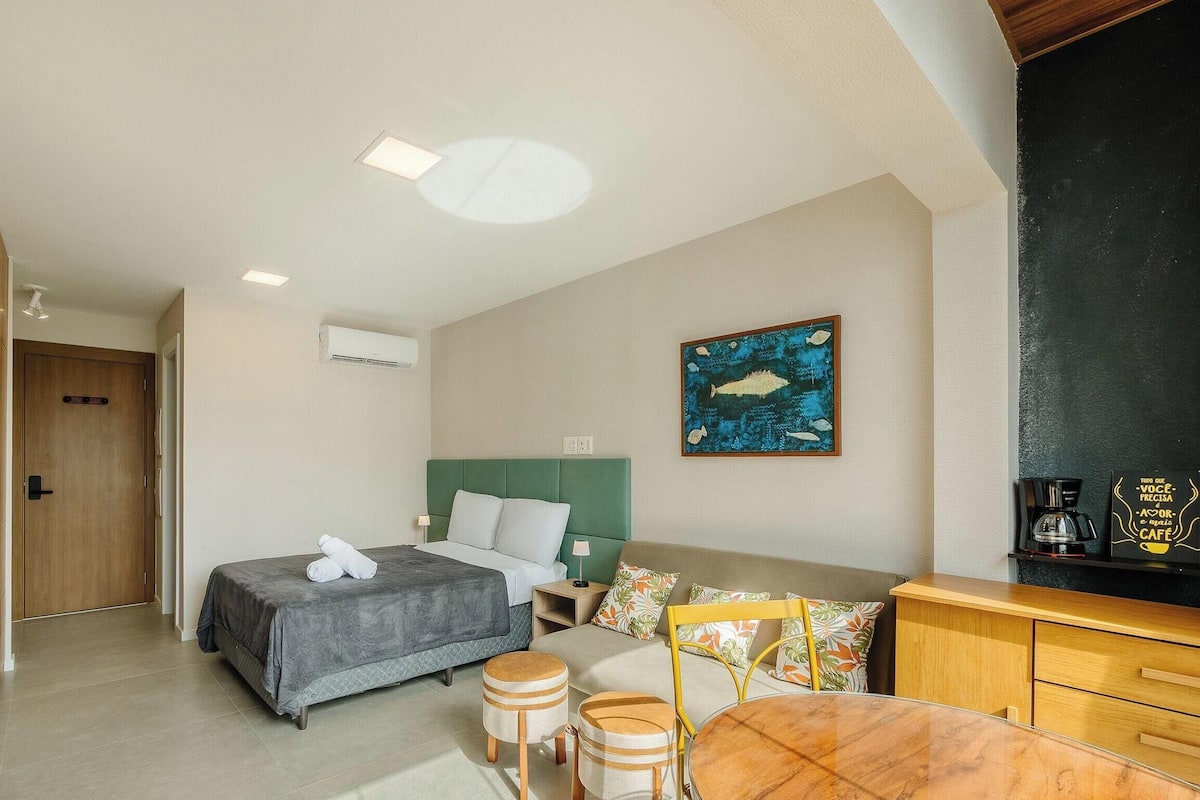 A hotel room with a bed, a small table, a chair, a sofa, a coffee machine, and an air conditioning unit.