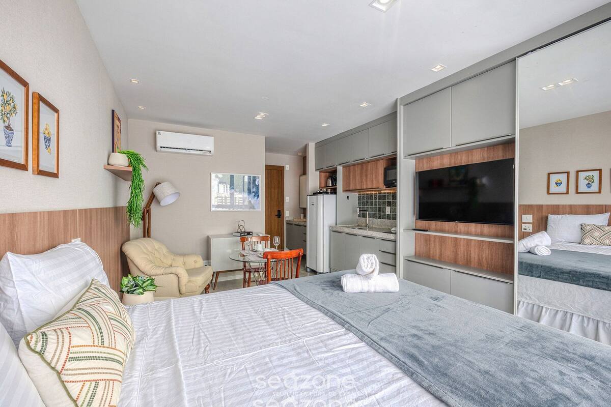 A modern hotel room with a bed, a desk, a chair, a TV, and a kitchenette.
