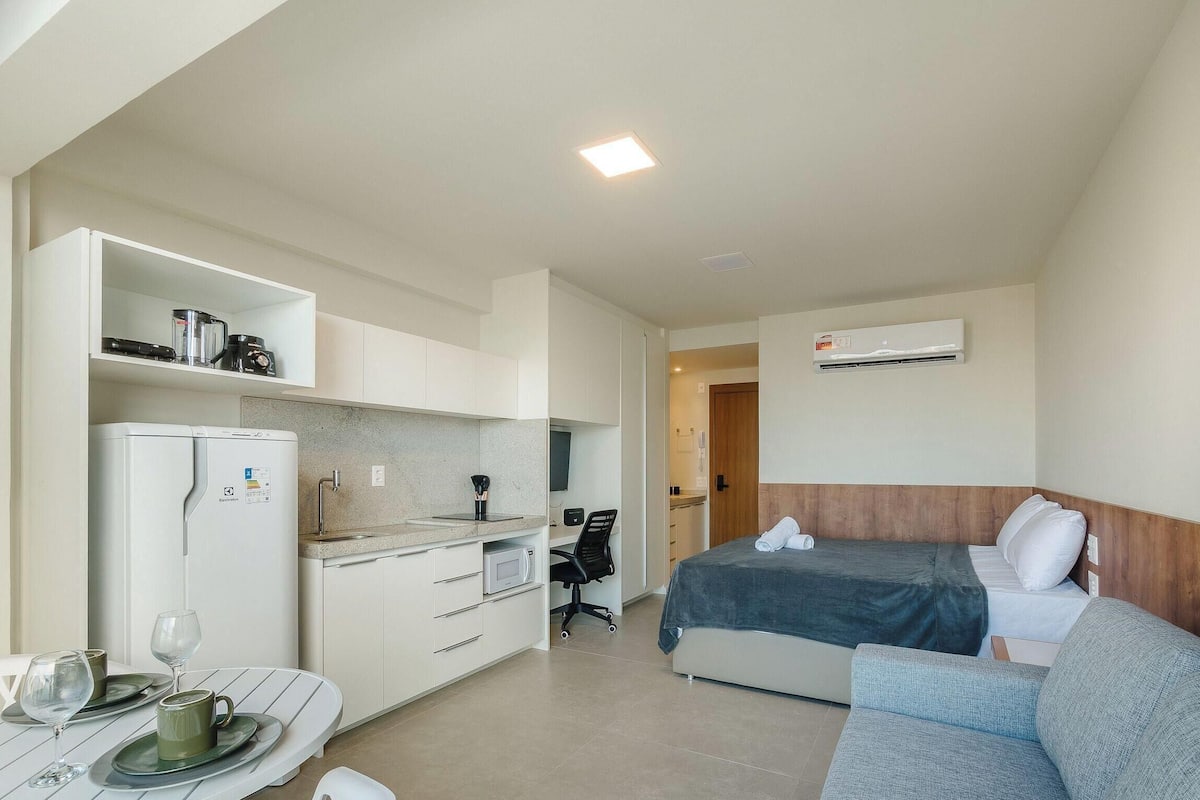 A compact hotel room with a kitchenette, a bed with a blue blanket, a grey sofa, and an air conditioning unit.