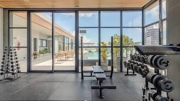 Un gimnasio con equipo de levantamiento de pesas, una banca y vistas a un patio exterior.