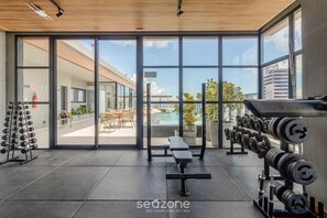 Un gimnàs amb equips de musculació, una panca i vista a una zona de pati exterior.