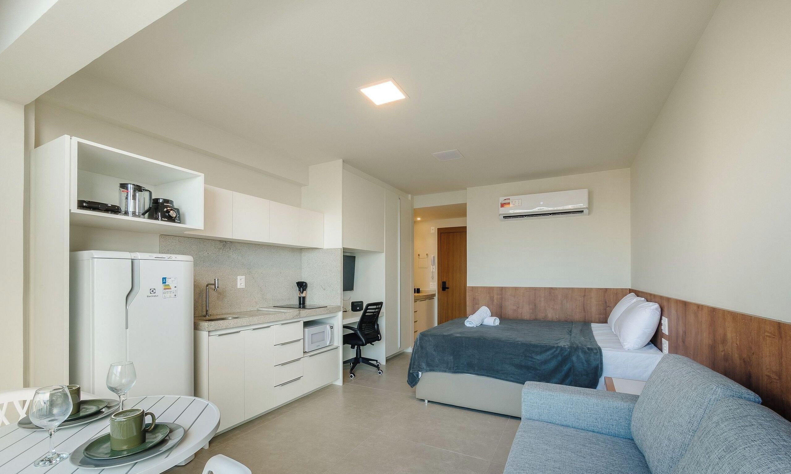 A compact hotel room with a kitchenette, a bed with a headboard, a seating area with a table and chairs, and an air conditioning unit.