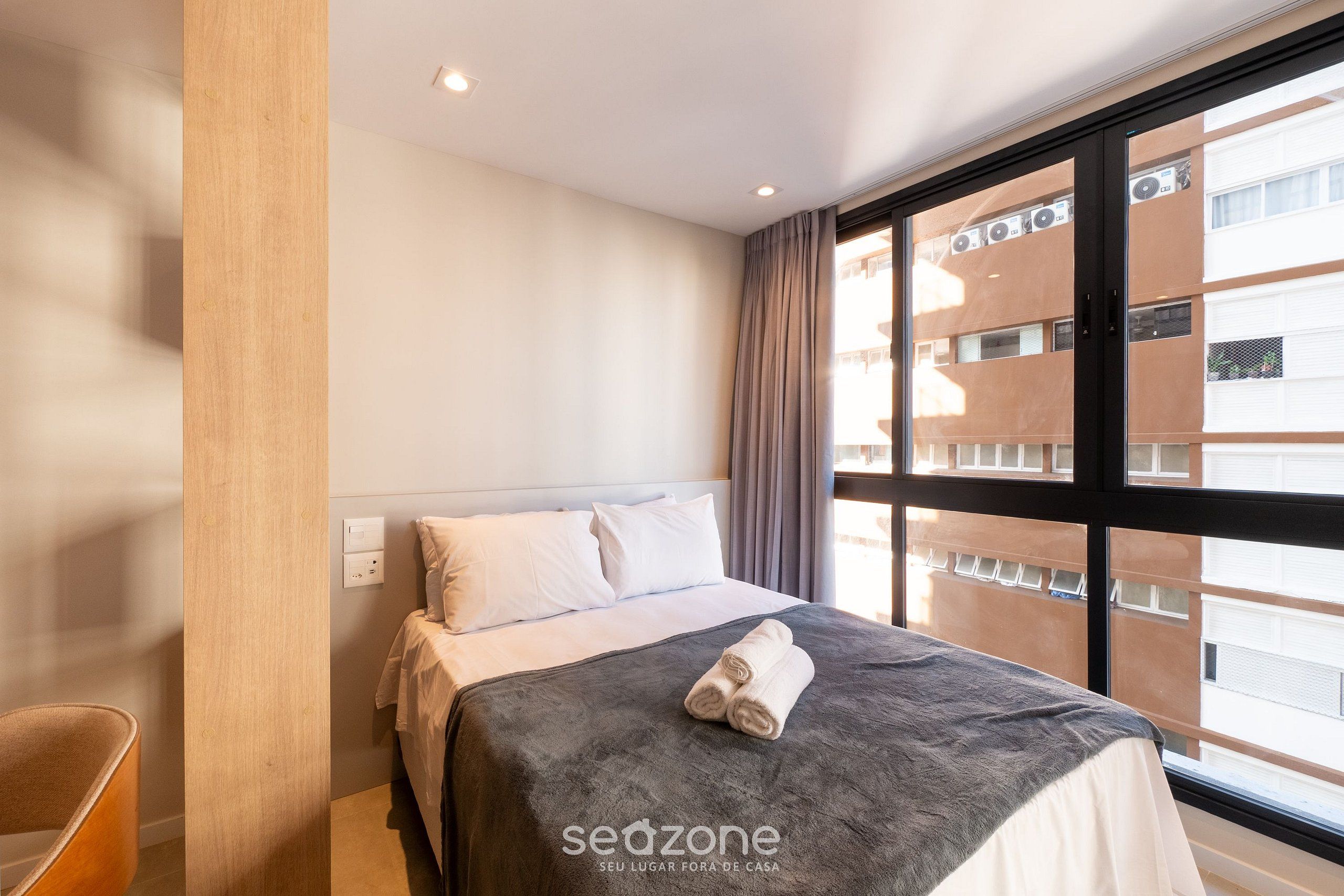 A hotel room with a bed, a window with curtains, a wooden column, and a view of a building.