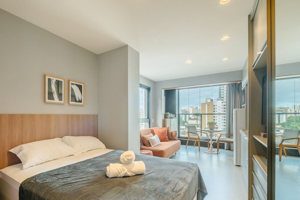 Habitación de hotel con cama, zona de estar con mesa y sillas, y un gran ventanal con vistas a la ciudad.