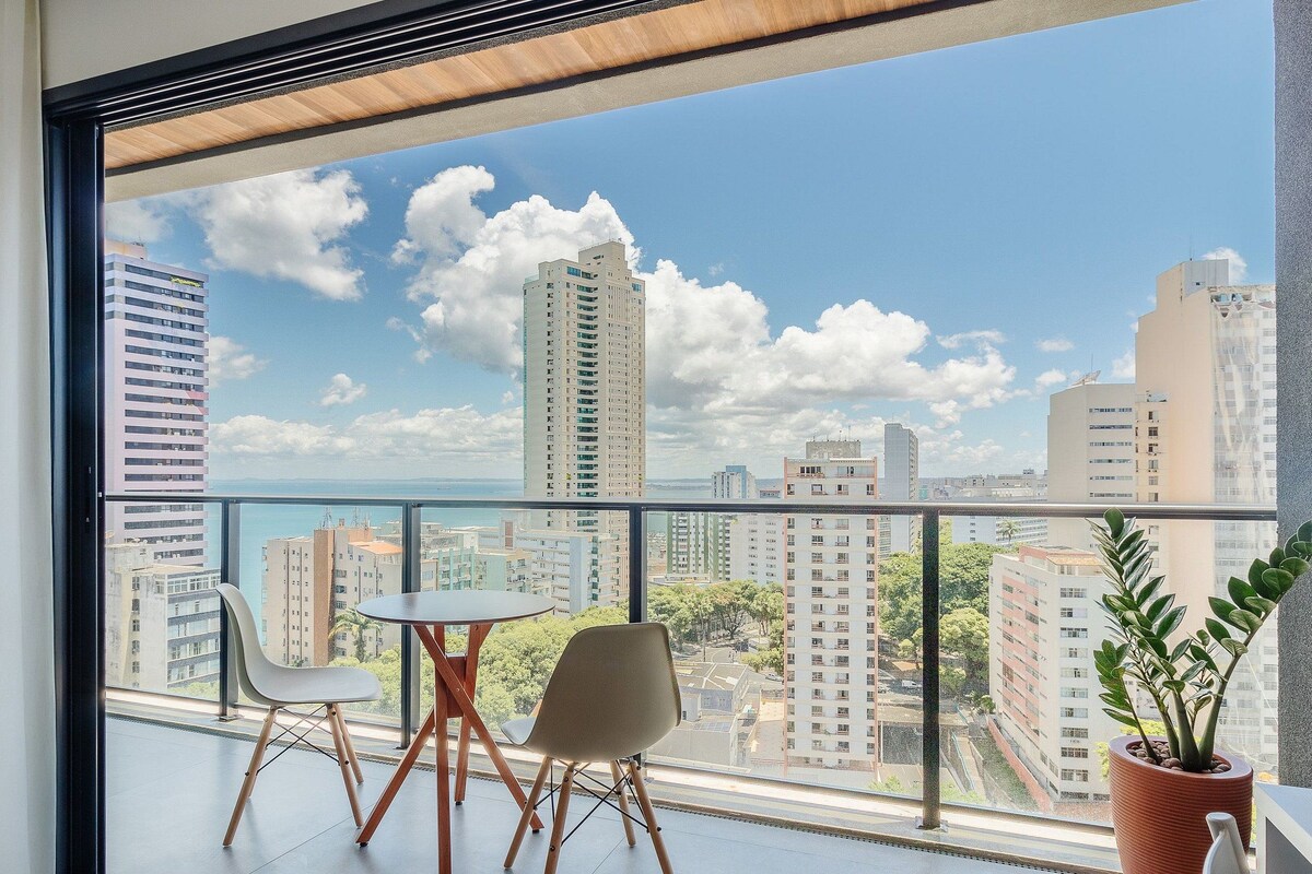 A balcony with a table and chairs, a potted plant, and a view of a cityscape with tall buildings.