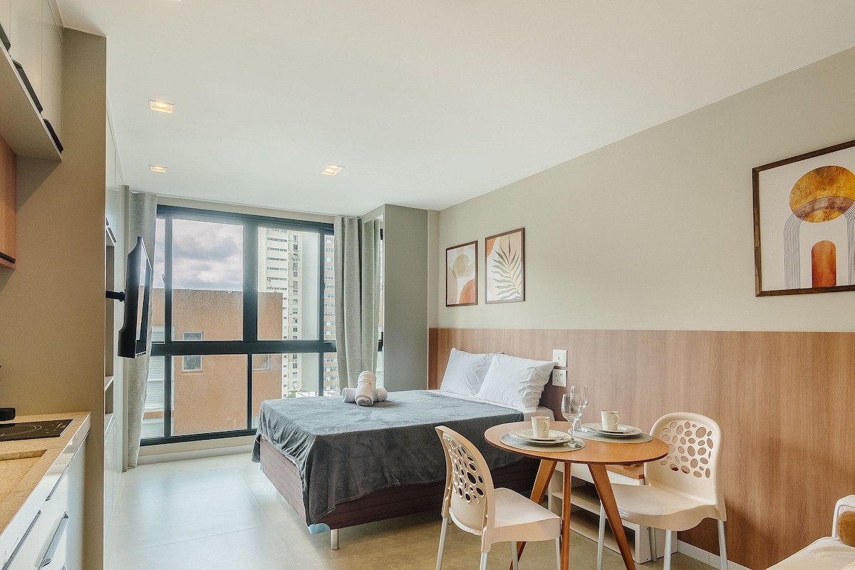A hotel room with a bed, a dining table with chairs, a TV, and a large window with a view of buildings.
