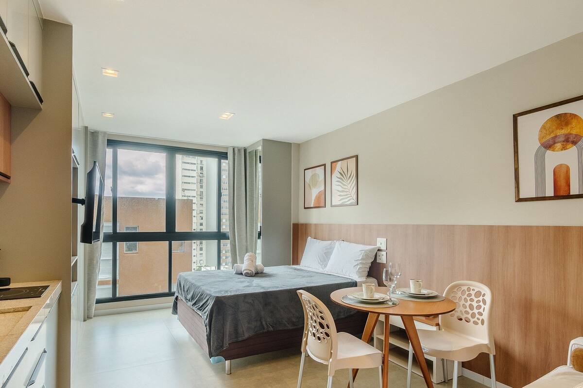 A hotel room with a bed, a dining table with chairs, a TV, and a large window with a view of buildings.