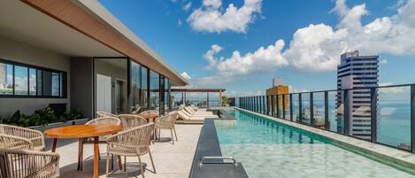 A rooftop pool area with lounge chairs and tables, a clear view of the ocean, and a modern building in the background.