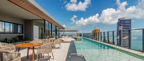 A rooftop pool area with lounge chairs and tables, a clear view of the ocean, and a modern building in the background.