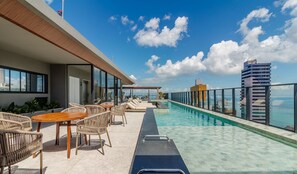 Ein Poolbereich auf dem Dach mit Liegestühlen und Tischen, freier Blick auf das Meer und ein modernes Gebäude im Hintergrund.
