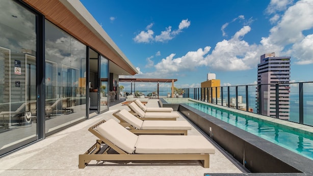 A rooftop pool with lounge chairs, a glass-enclosed area, and a view of the ocean and a tall building. - Stay With Style and Convenience in Salvador LIV (Salvador)