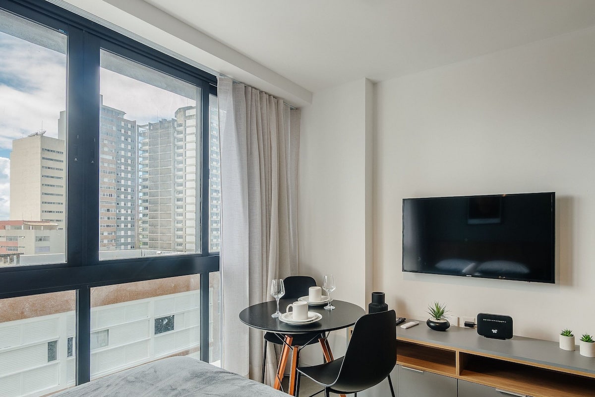 A modern hotel room with a balcony, a dining area, a flat-screen TV, and a small plant.