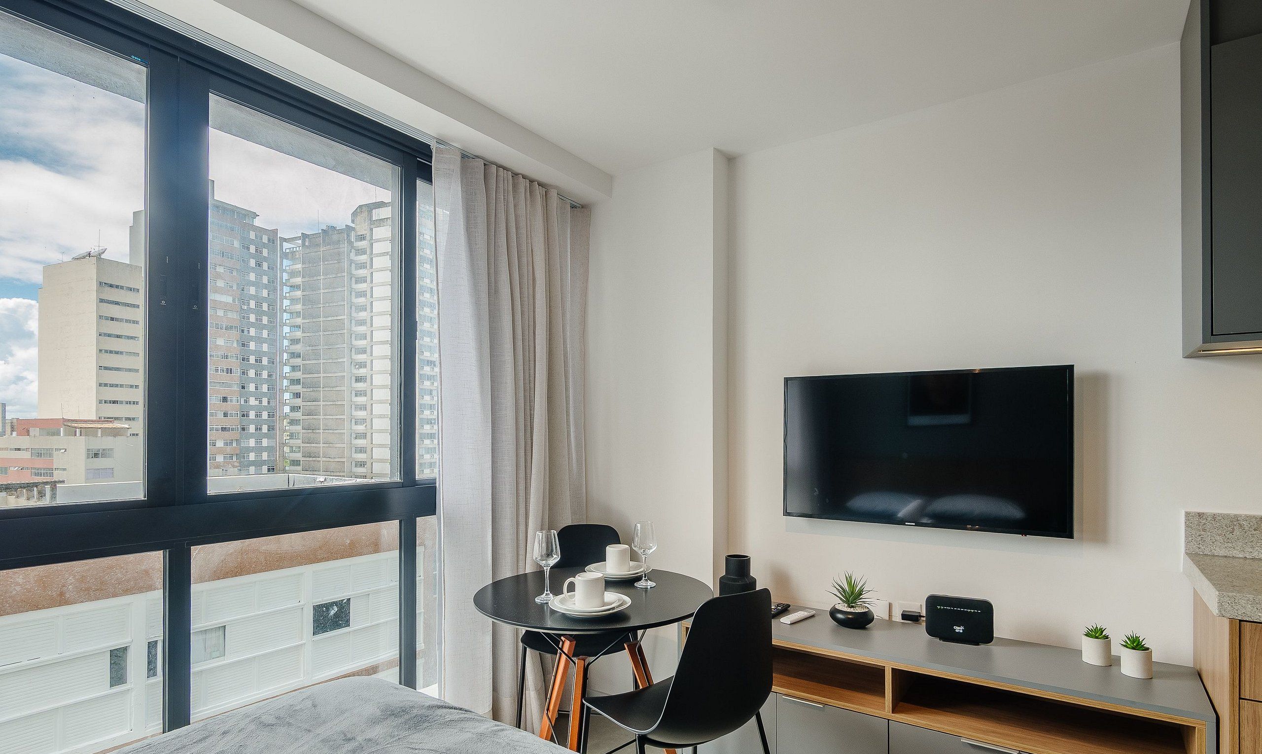 Quarto de hotel moderno com varanda, área de refeições, televisão de tela plana e uma pequena planta.