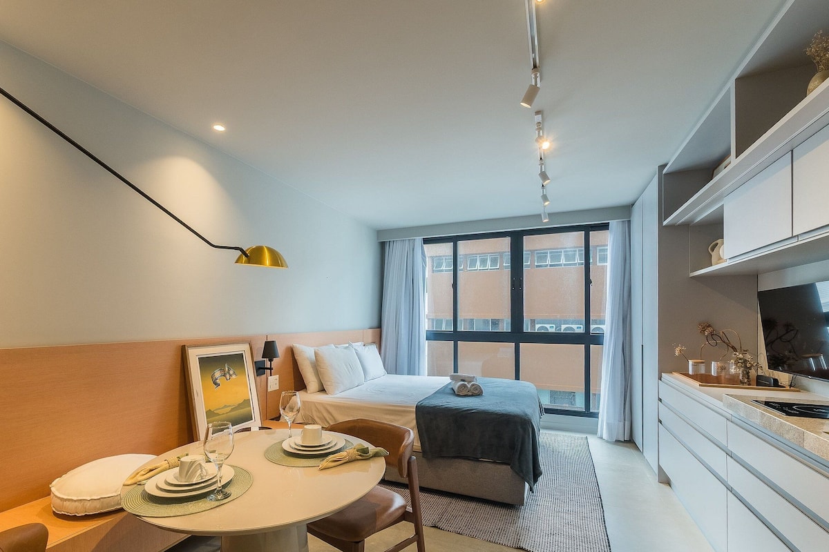 A compact hotel room with a bed, a dining table with a chair, a small kitchen area, and a large window providing a view of the outside.