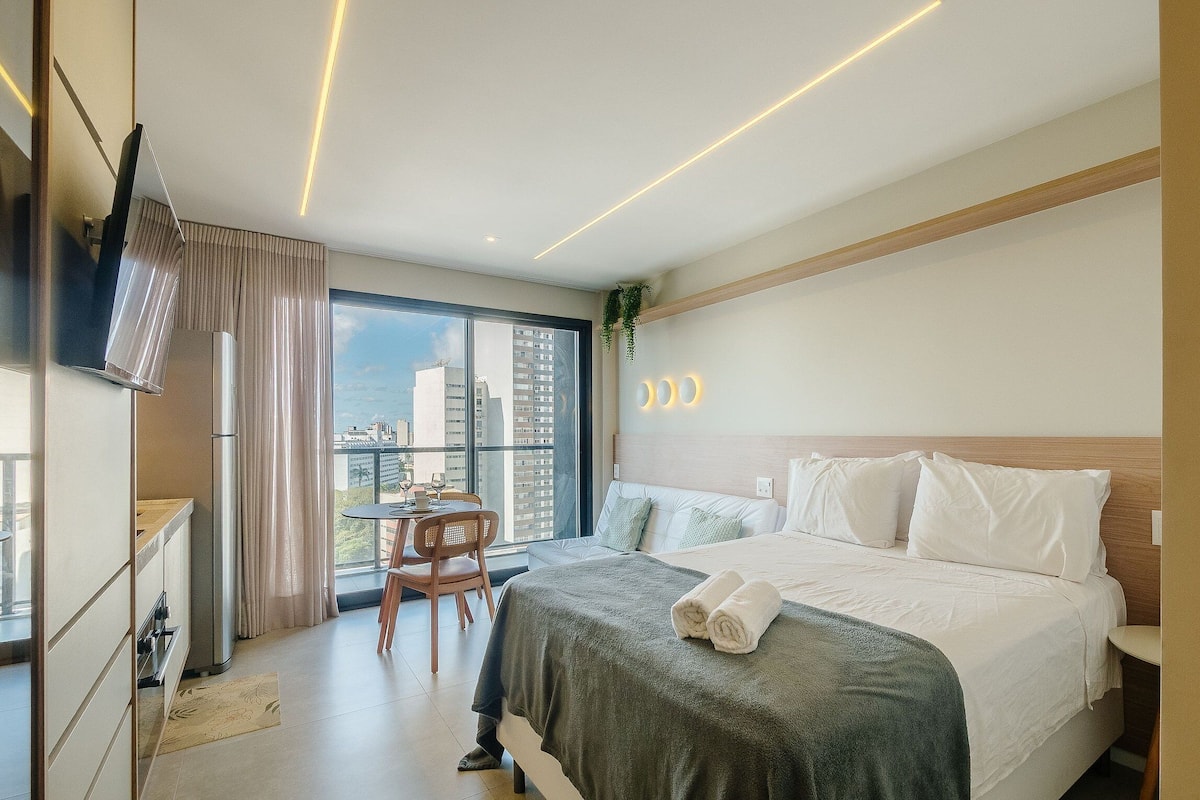 A hotel room with a bed, a small table with chairs, a TV, and a large window with a city view.