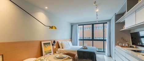 Una habitación de hotel compacta con cama, una mesa de comedor con una silla, una pequeña cocina y un amplio ventanal con vistas al exterior.