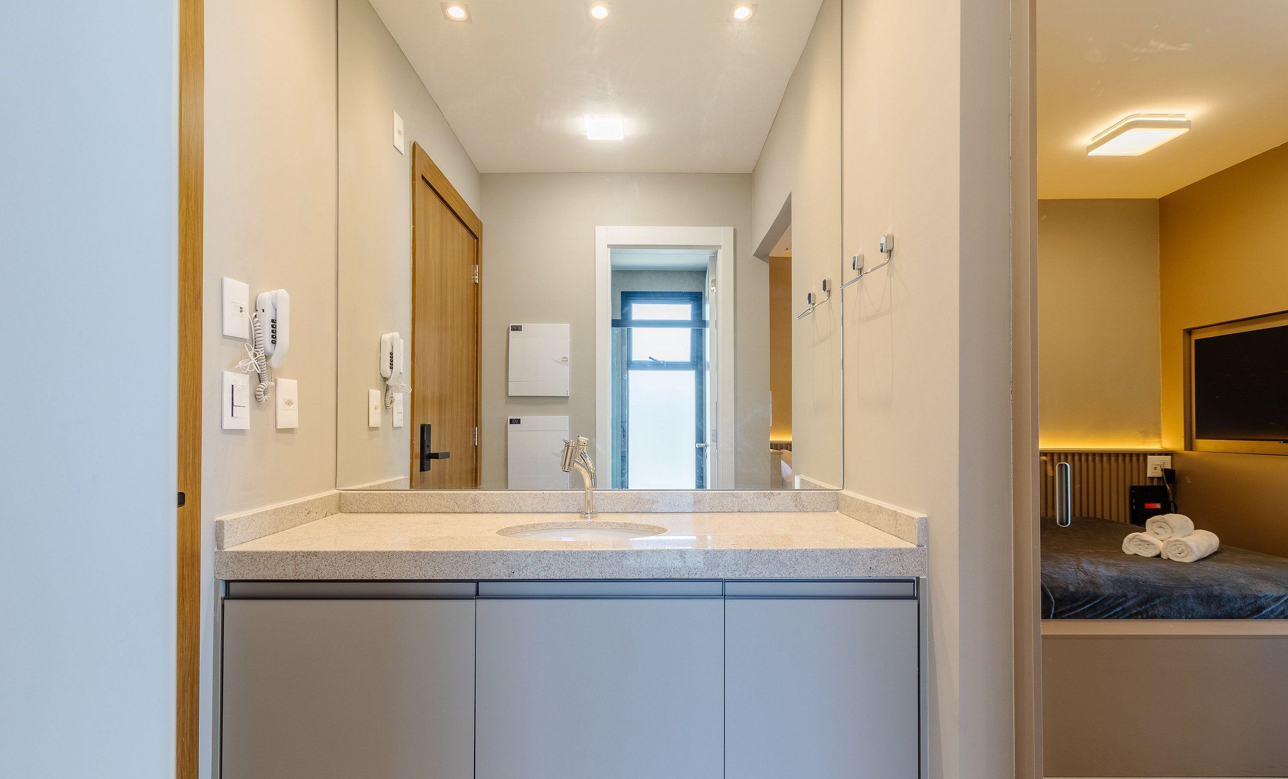 A modern bathroom with a large mirror, a sink, and a door leading to a bedroom.
