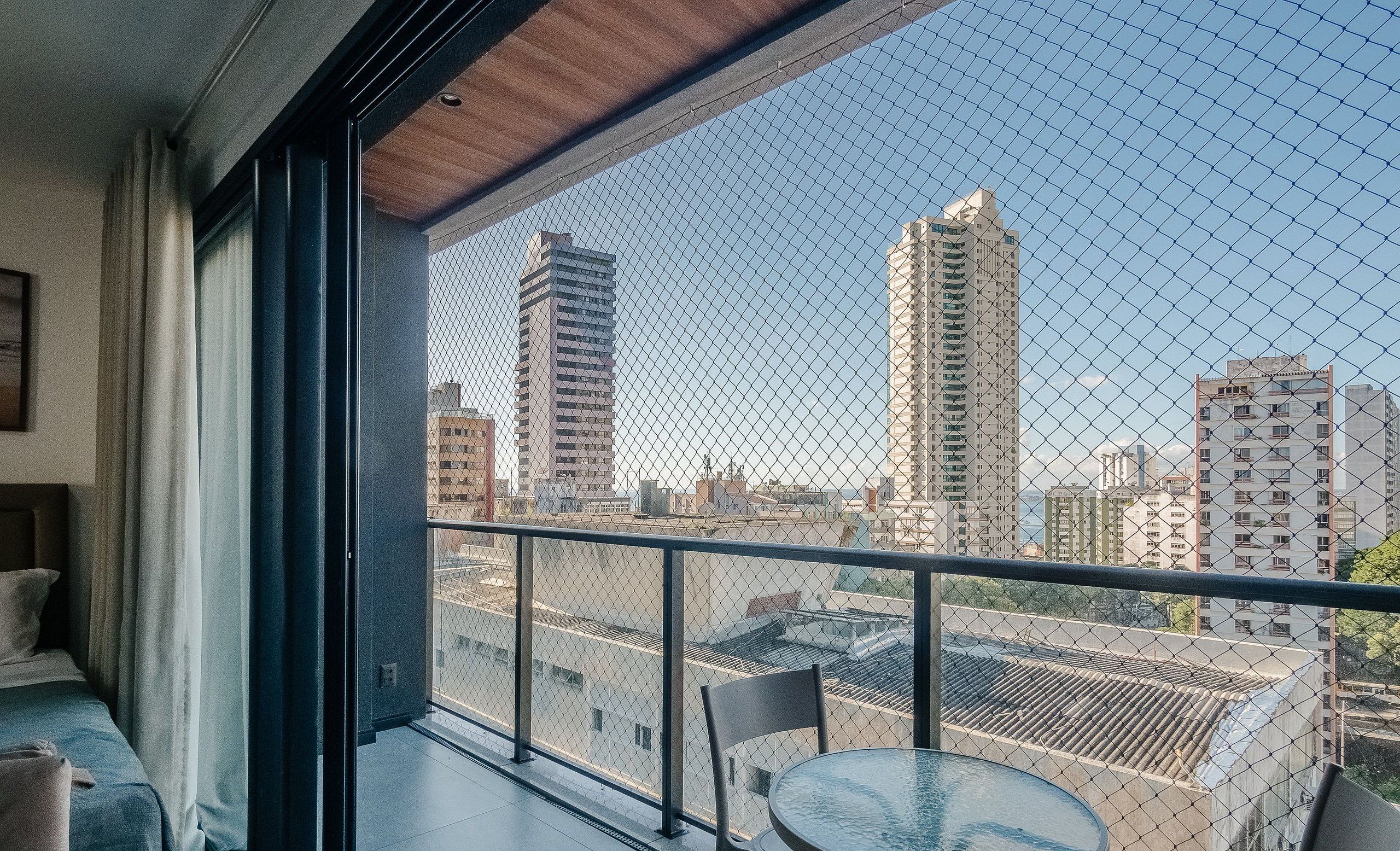 Un balcón con una cerca de malla metálica, una mesa y sillas, con vista a un paisaje urbano de edificios altos.