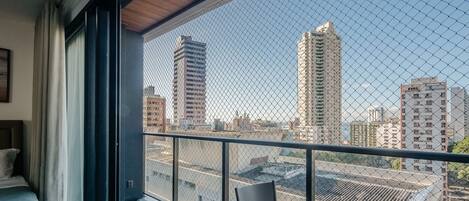 Un balcón con una cerca de malla metálica, una mesa y sillas, con vista a un paisaje urbano de edificios altos.
