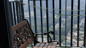 Un balcon doté d’un banc en bois et d’un siège rembourré, offrant une vue sur un paysage urbain avec de hauts immeubles.