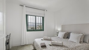 Ein Schlafzimmer mit Bett, Kissen, einem Nachttisch, einem Sessel, einem Fenster mit Blick und einem Vorhang.