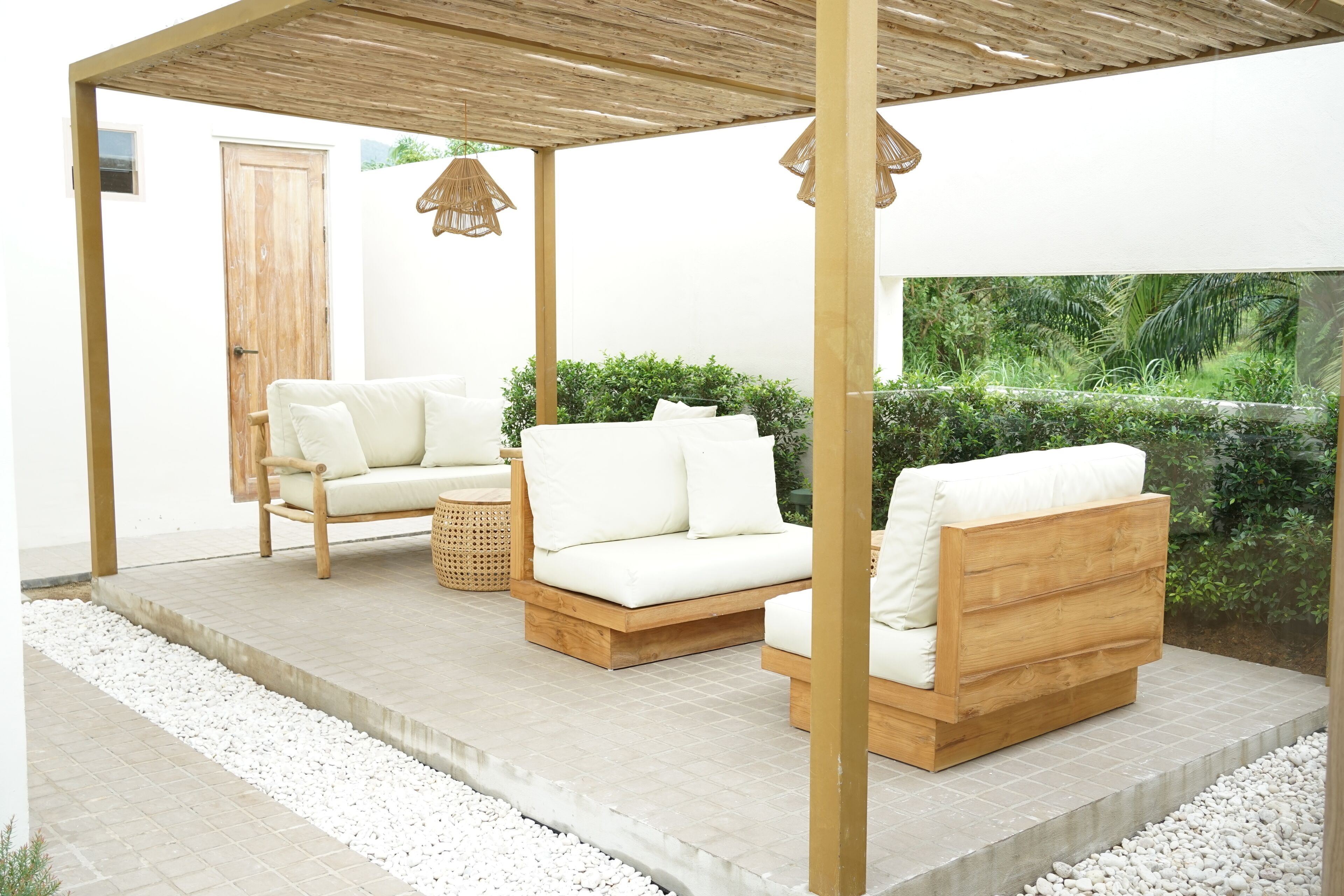 A covered outdoor seating area with two sofas and a chair, a wooden door, and a view of greenery.
