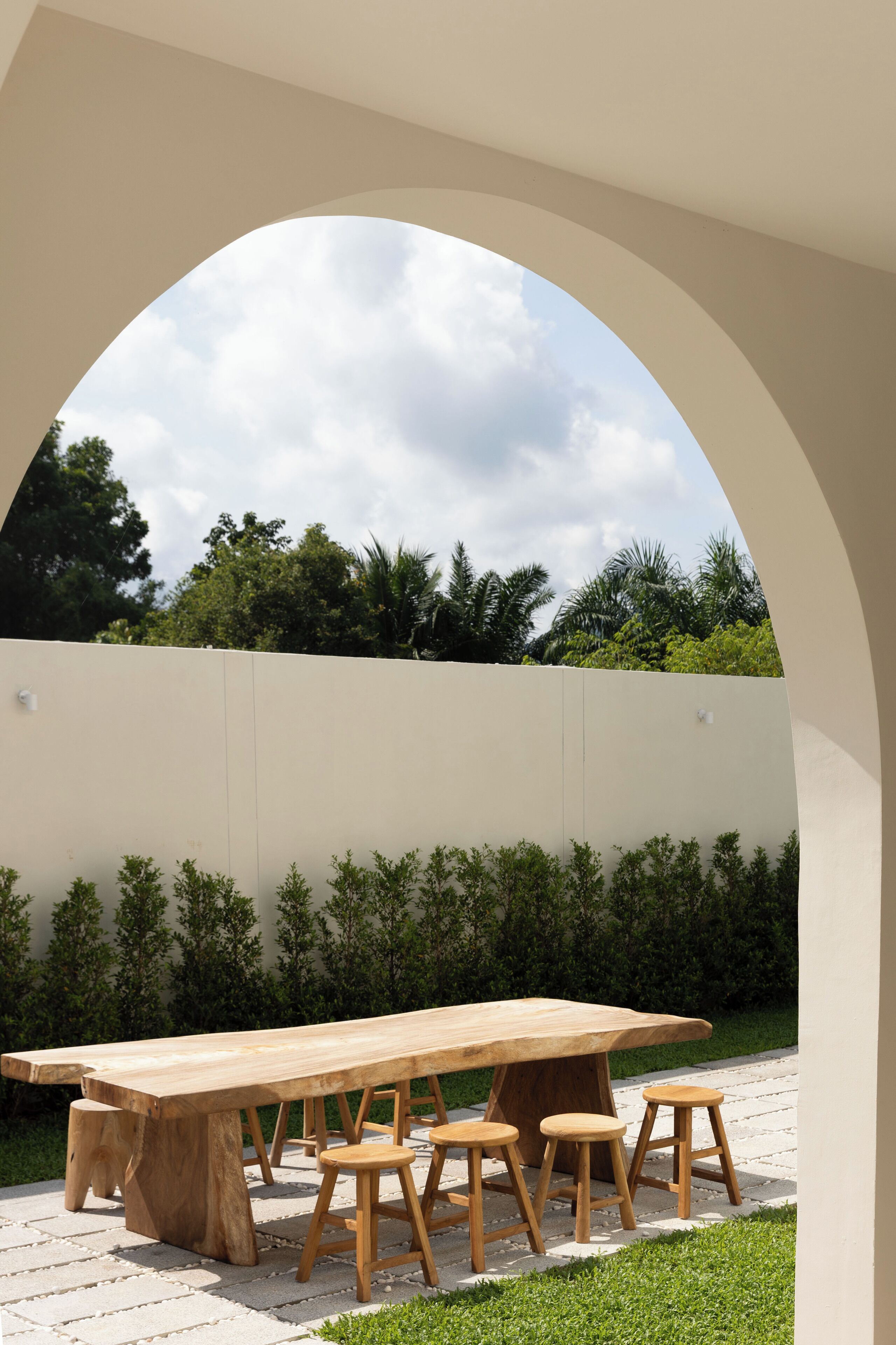 An outdoor dining area with a large wooden table and stools, set against a white wall with greenery in the background.