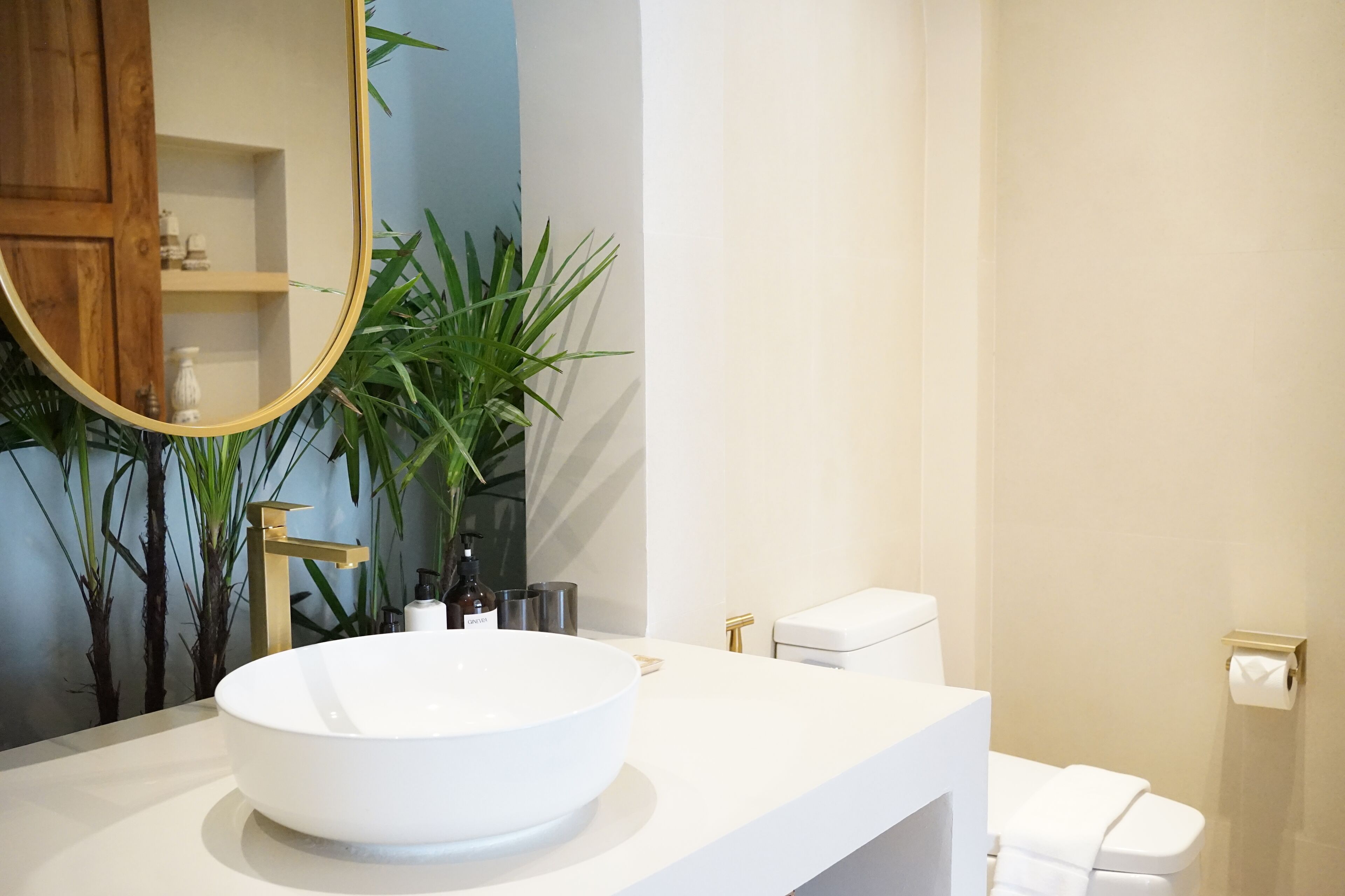 Een badkamer met een witte wastafel, een gouden kraan en een spiegel. Er is een toilet en een plant in de hoek.