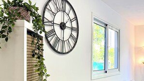 A living room with a large wall clock, a sofa, a small side table with a plant, and a window with a view of greenery.