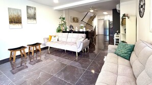 A spacious living area with a sofa, two wooden stools, a dining table, and a kitchen in the background.