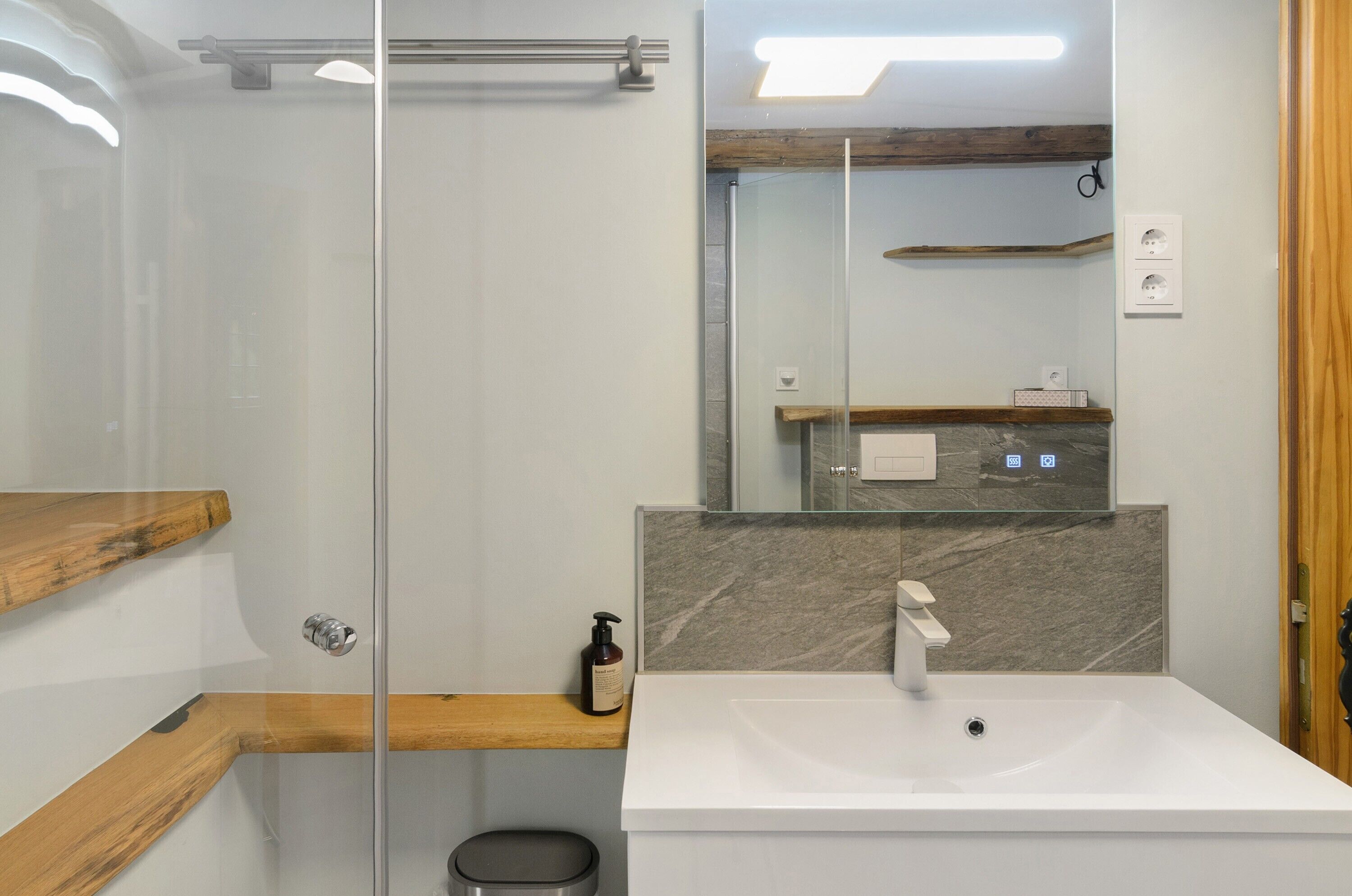 Un baño moderno con cabina de ducha de vidrio, estante de madera, salpicadero de mármol y lavamanos blanco.