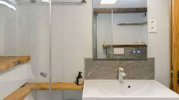Un baño moderno con cabina de ducha de vidrio, estante de madera, salpicadero de mármol y lavamanos blanco.