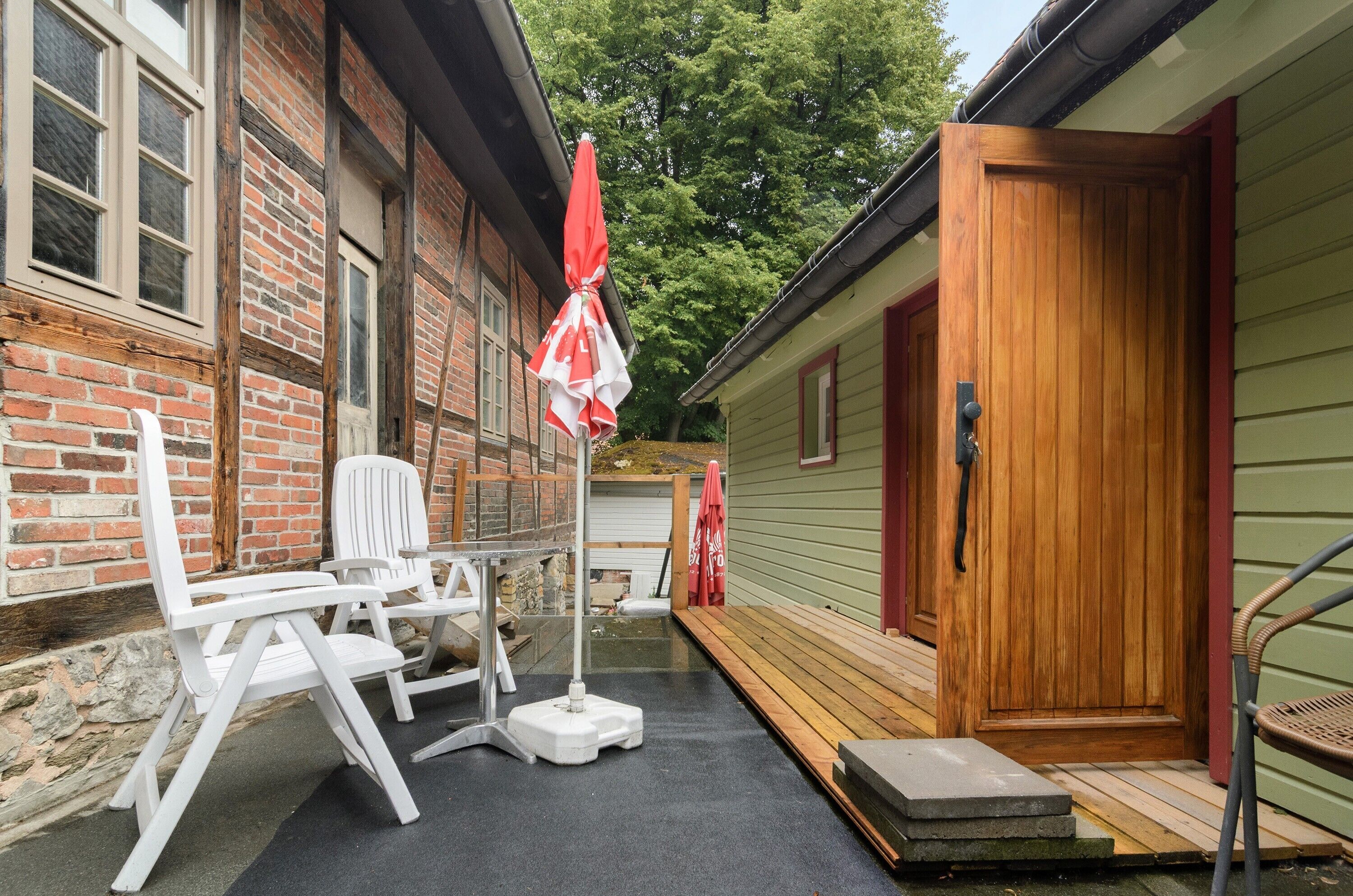 Eine kleine, rustikale Holzhütte mit Holztür, Ziegelaußenwand und einer kleinen Veranda mit weißen Stühlen und einem roten Sonnenschirm.