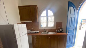 A kitchen with wooden cabinets, a white refrigerator, a window with curtains, and a blue door. - Aeolos Apartments (Santorini)