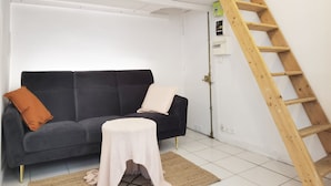 Une pièce avec un canapé gris foncé, un tabouret blanc en tricot, une échelle en bois et un plafond blanc avec des poutres en bois.