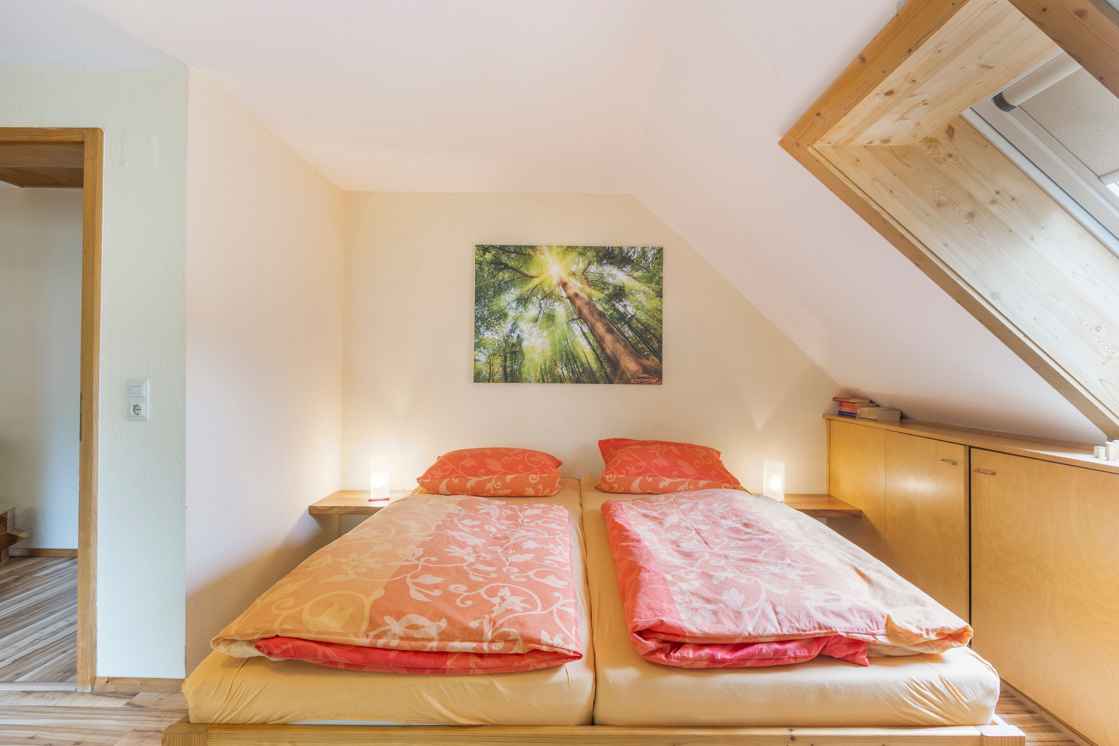 Ein Schlafzimmer mit schrägemaßtem Dach, einem Bett mit floralem Bettzeug, einem Nachttisch und einem an der Wand hängenden Bild.