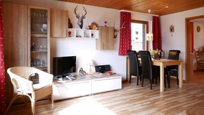Ein gemütliches Wohnzimmer mit Holzbalkendecke, einem Fernseher, einer Couch, einem Esstisch mit Stühlen und einer Vitrine.